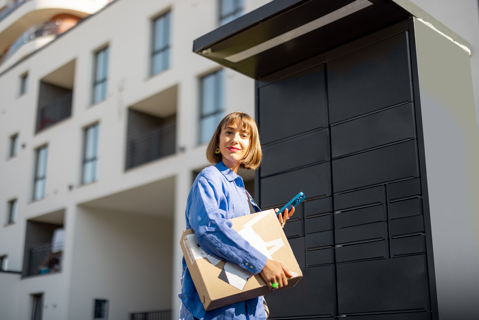 Woman with phone and parcel near automatic post office machine in residential district